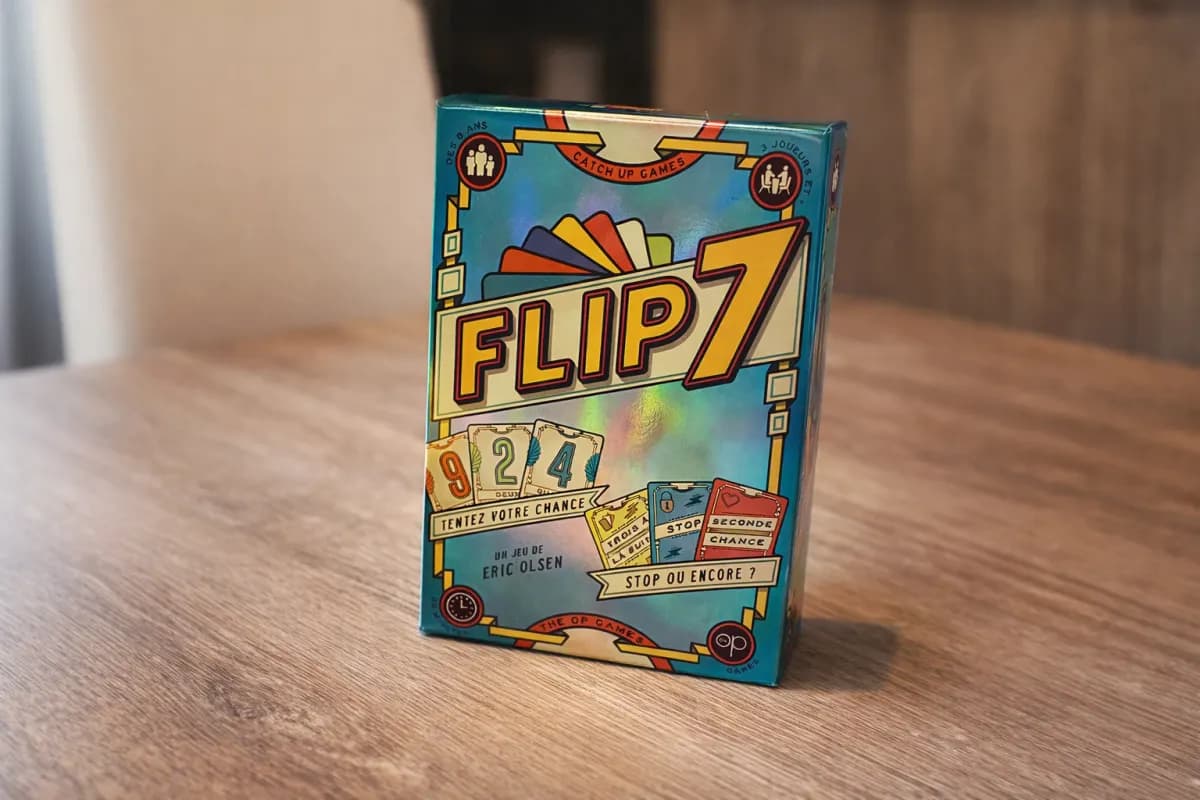 Boîte colorée du jeu de cartes Flip 7 posée debout sur une table en bois, avec un arrière-plan intérieur flou.